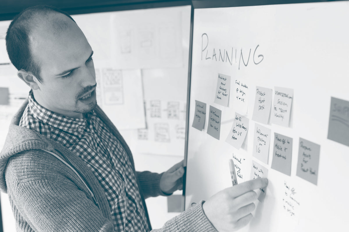 A man adds another sticky note to a group of notes already on a whiteboard. He is about to say something and is wearing a checked shirt and hooded cardigan. 