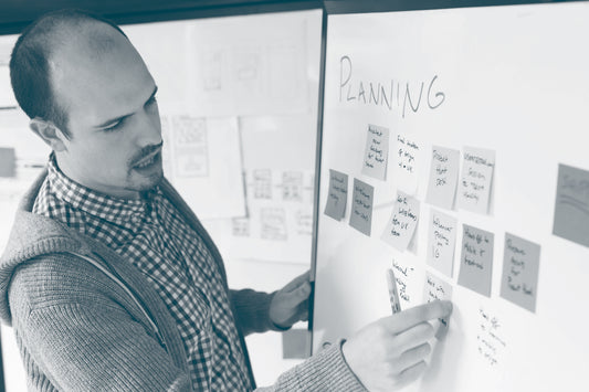 A man adds another sticky note to a group of notes already on a whiteboard. He is about to say something and is wearing a checked shirt and hooded cardigan. 
