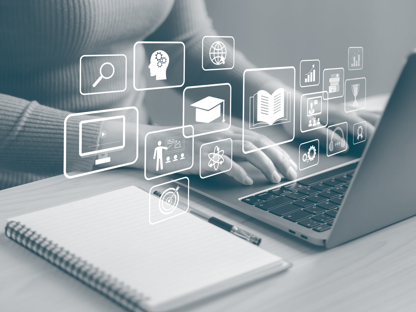 A person in a sweater types on a laptop at a desk, with a notebook and pen in the foreground and white digital icons floating above the screen: they represent scholarship, digital and print dissemination, metrics and process.