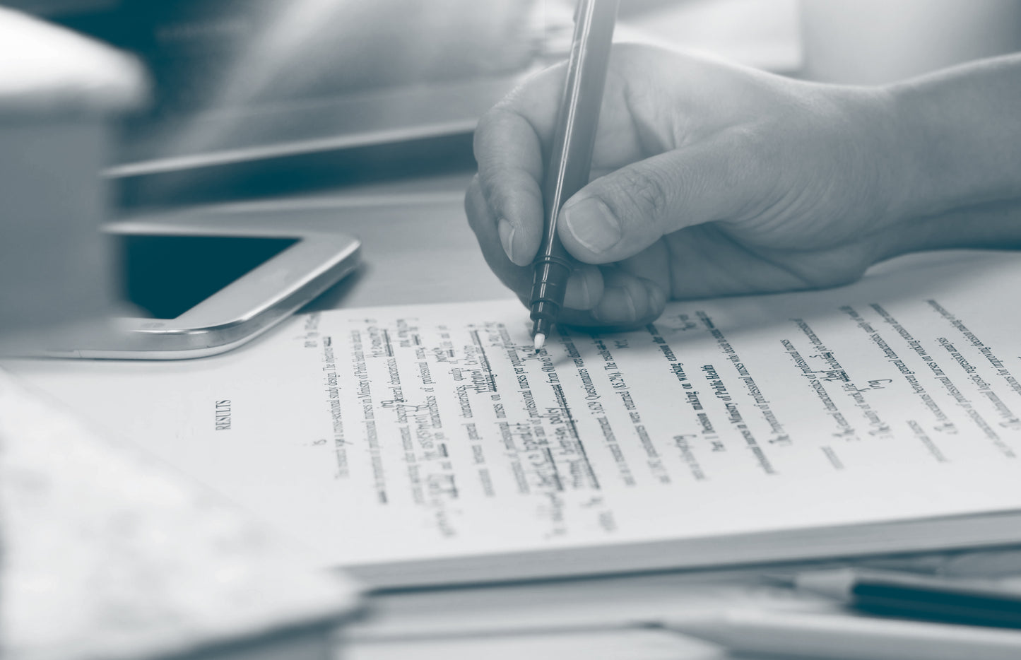 A close-up of a hand holding a pen and marking up text on a printed document on a desk, with a smartphone and other desk items blurred in the background.