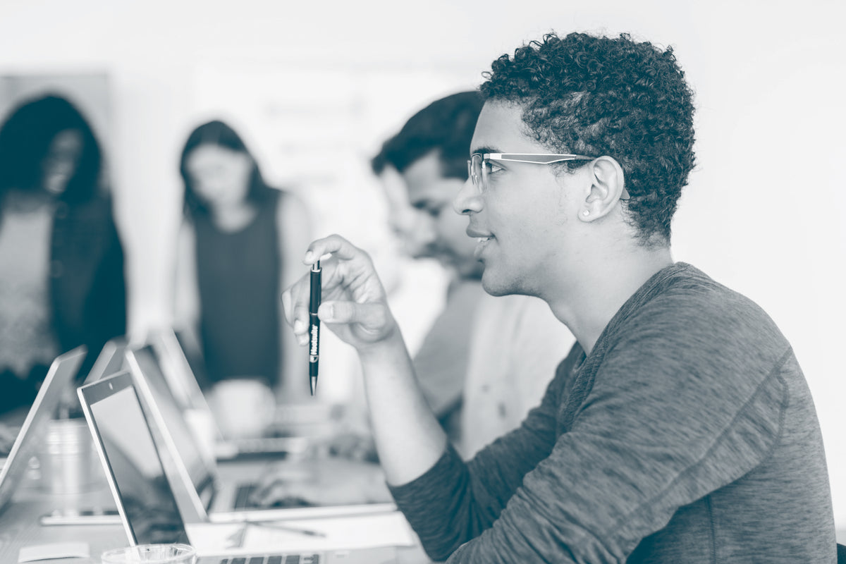 A main in a group setting holds a pen and sits in front of an open laptop. He is engaging with the person opposite (who is out of shot for us). In the background, we can see others in the group sitting or standing and engaging with each other or their work.