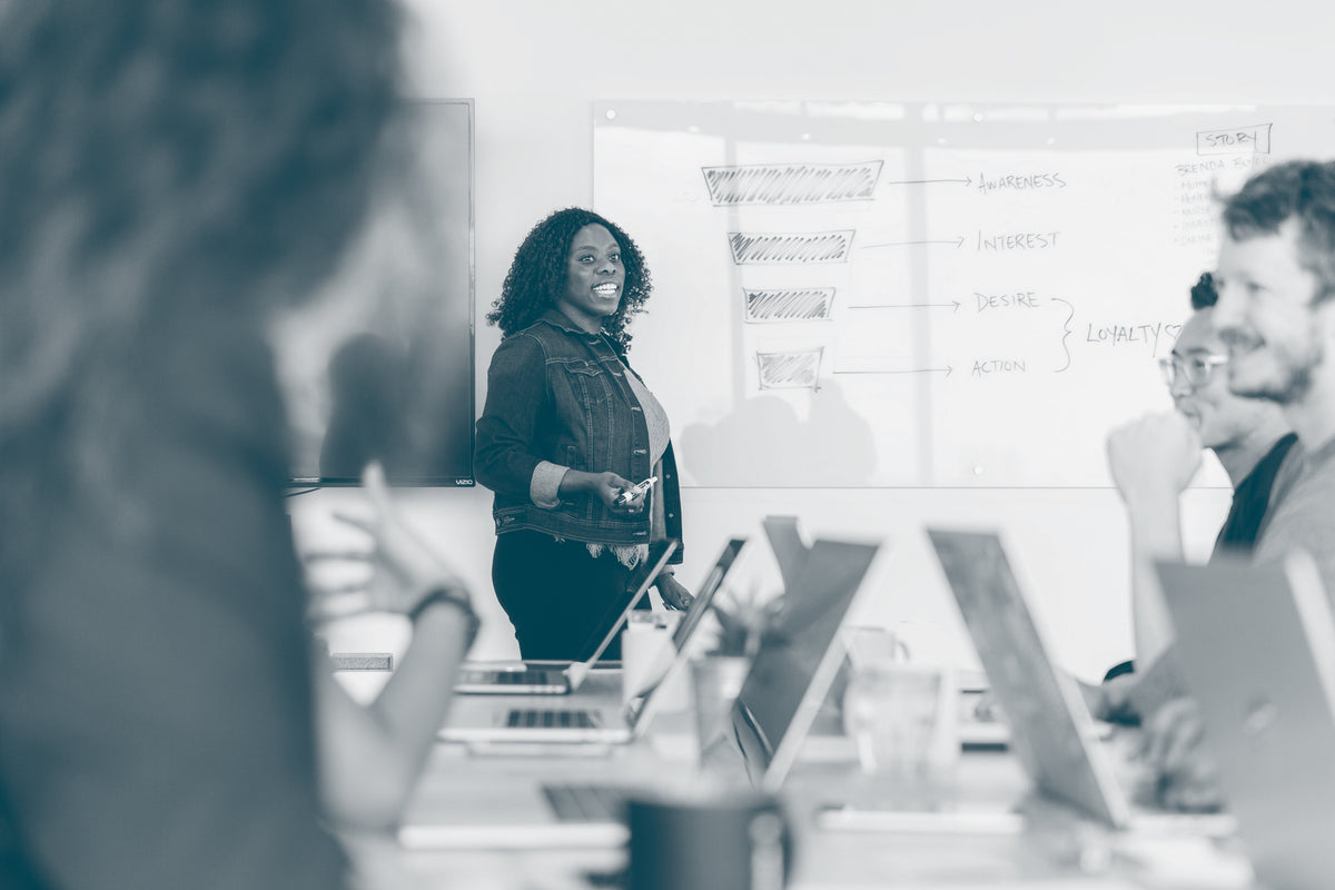 A women is leading an office-based training session. She is standing in front of a whiteboard where she has been writing and is talking to the group of seated attendees. They are engaged and smiling, with their laptops in front of them on the table.