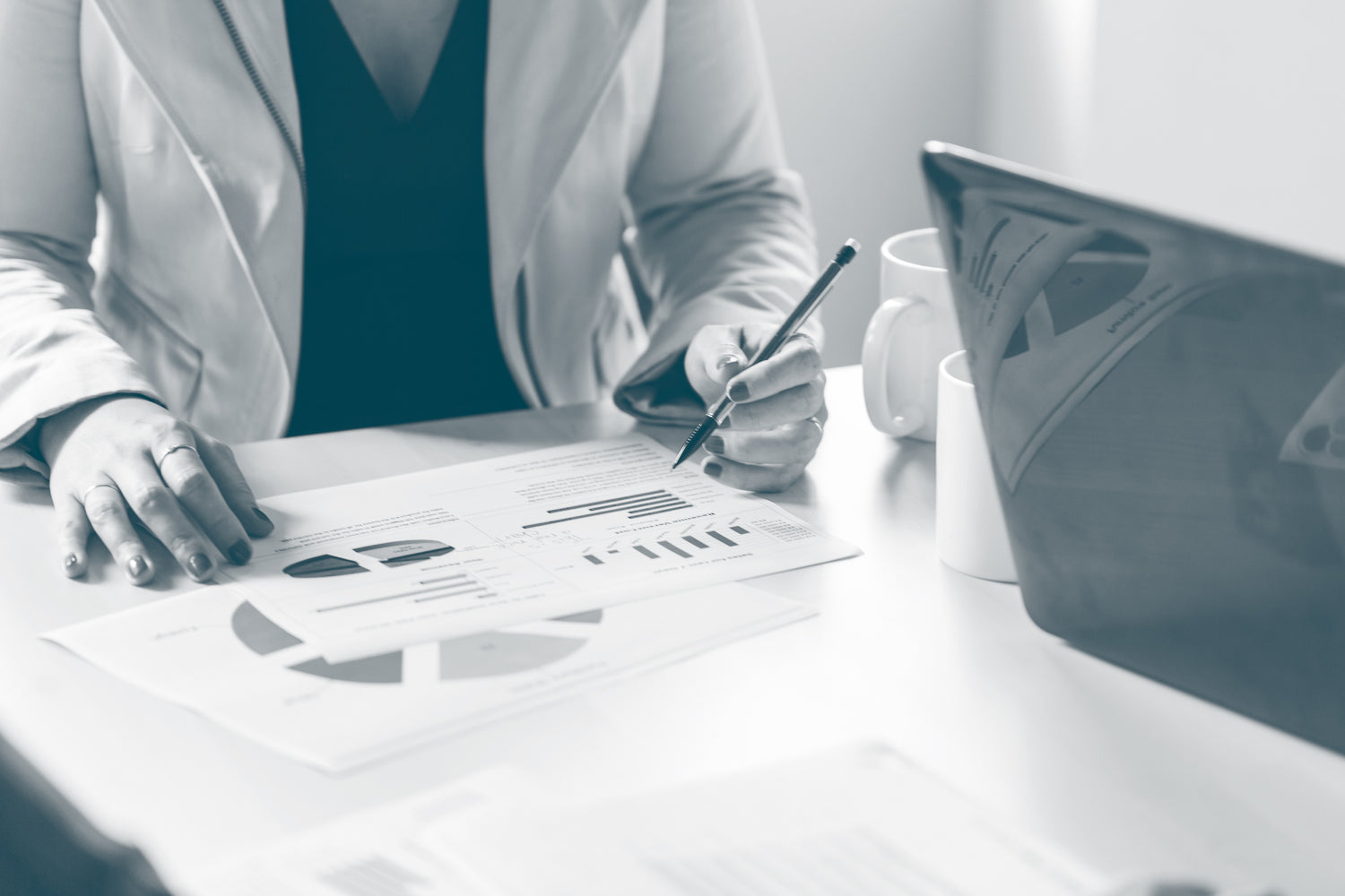 A woman sits at a desk, holding a pen, while looking through printed data and reporting. An open laptop and white mugs also sit on the desk.
