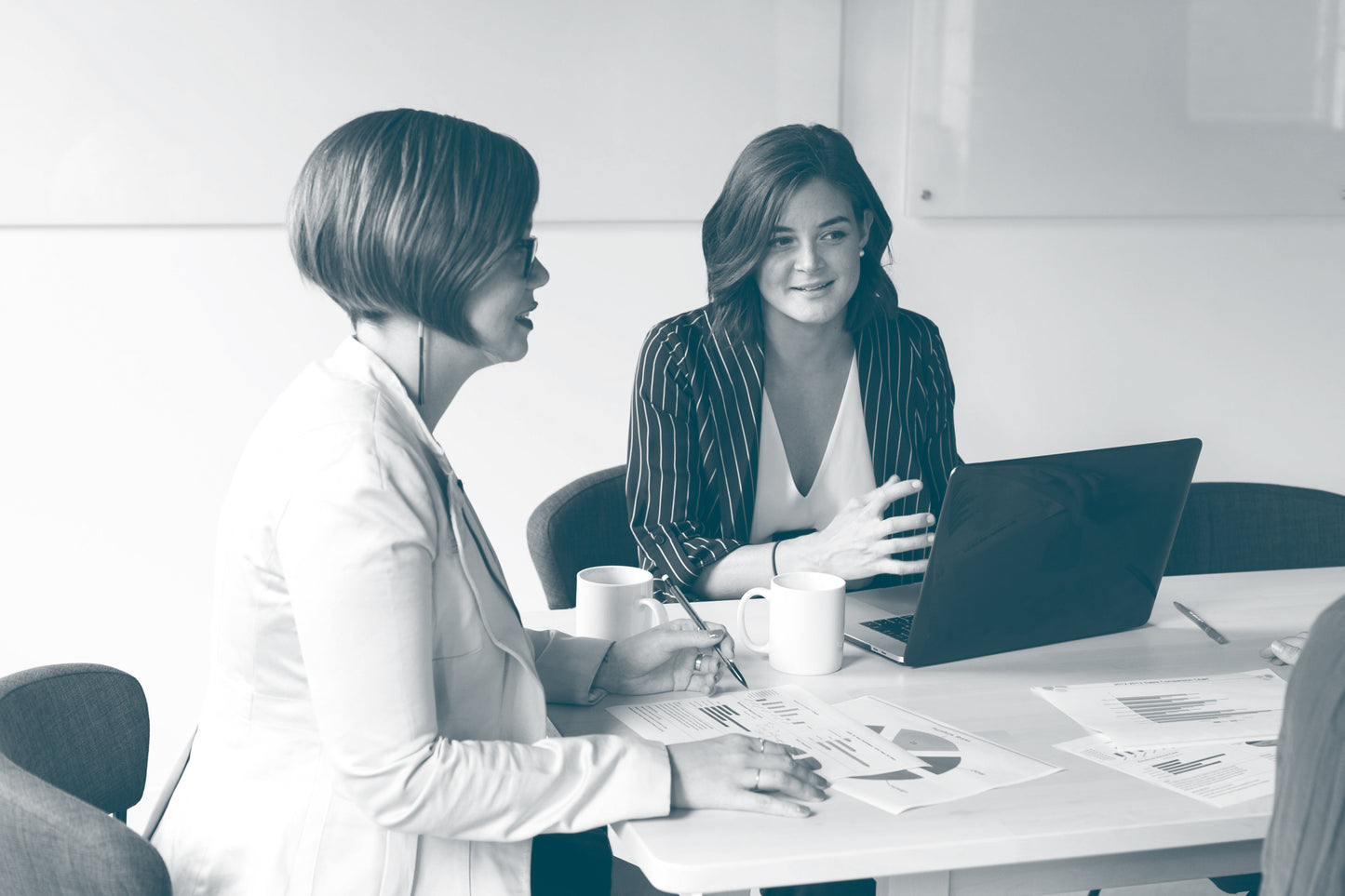 Two business women are in conversation, sitting at stable with an open laptop and printed reports including data.